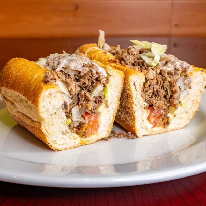 Two halves of a cheesesteak sandwich on a white plate, filled with meat, cheese, and vegetables.
