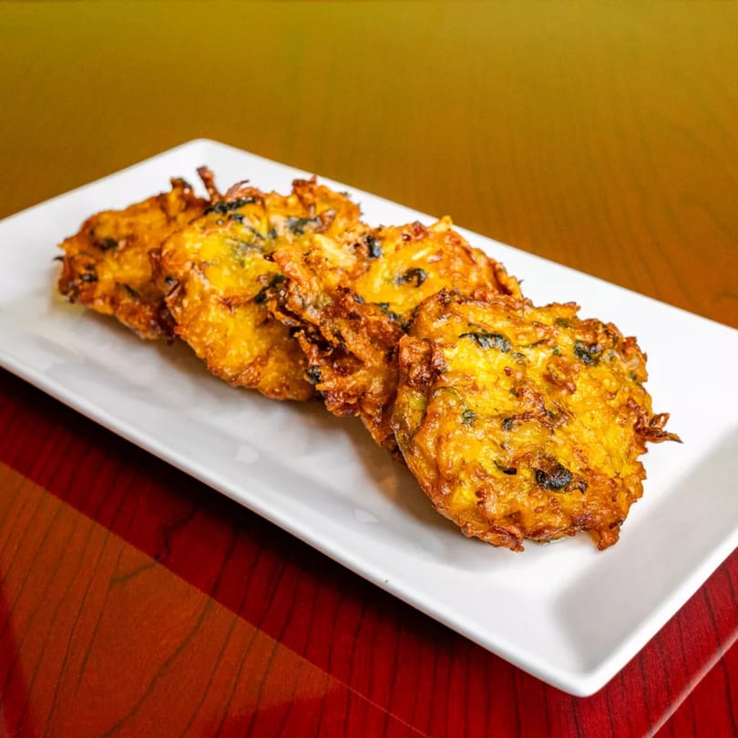 Four golden-brown, crispy fritters with dark specks, served on a long white rectangular plate.